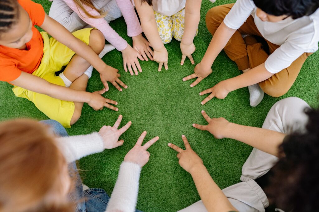 Paillettes et Arc-en-Ciel - vers une éducation coopérative et des enfants épanouis - Image montrant un groupe d'enfant assis en tailleur, en cercle au sol. Ils font un jeu ensemble avec leurs doigts. Cela représente la collaboration et la coopération.