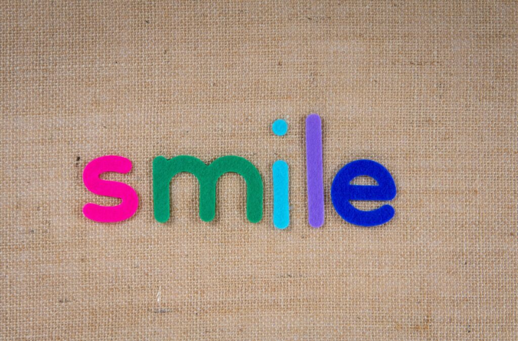 Paillettes et Arc-en-Ciel - vers une éducation coopérative et des enfants épanouis - Image de lettres en feutrine colorée, représentant le mot SMILE (sourire en français). Les lettres sont posées sur un morceau de toile de jute.